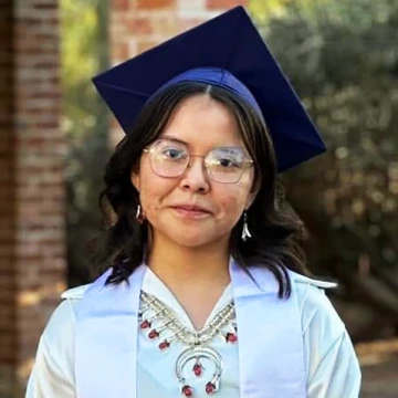 Michaela Hollowhorn poses in a graduation cap and white stole in front of two brick columns and greenery