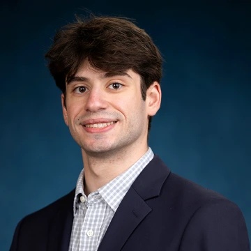 Spencer Metz smiles in a collared shirt and black jacket in a studio portrait with a dark blue background