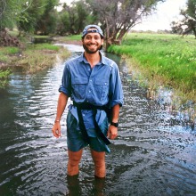 Josh Schachter wades trough a stream in shorts, long-sleeve denim shirt and backward hat.