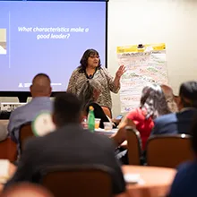 NNI Executive Director Joan Timeche address a crowd. A slide projected behind her reads "What characteristics make a good leader?"