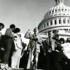 Morris K. Udall on the Capital steps behind a microphone with one first raised in the air. A crowd around him listens as he speaks.