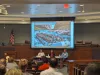 3 panelists sit under a projection screen with an AI-generated image of an enormous river raft full of business professionals floating down the Colorado River.