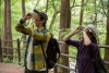 A man and woman look through binoculars on a forest path lined by a wooden fence.